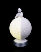 Bright Phases – Table Lamp showing a half illuminated textured moon on a round base with a small astronaut figure on top by 3DPRINTSWA for Lamps/Lights