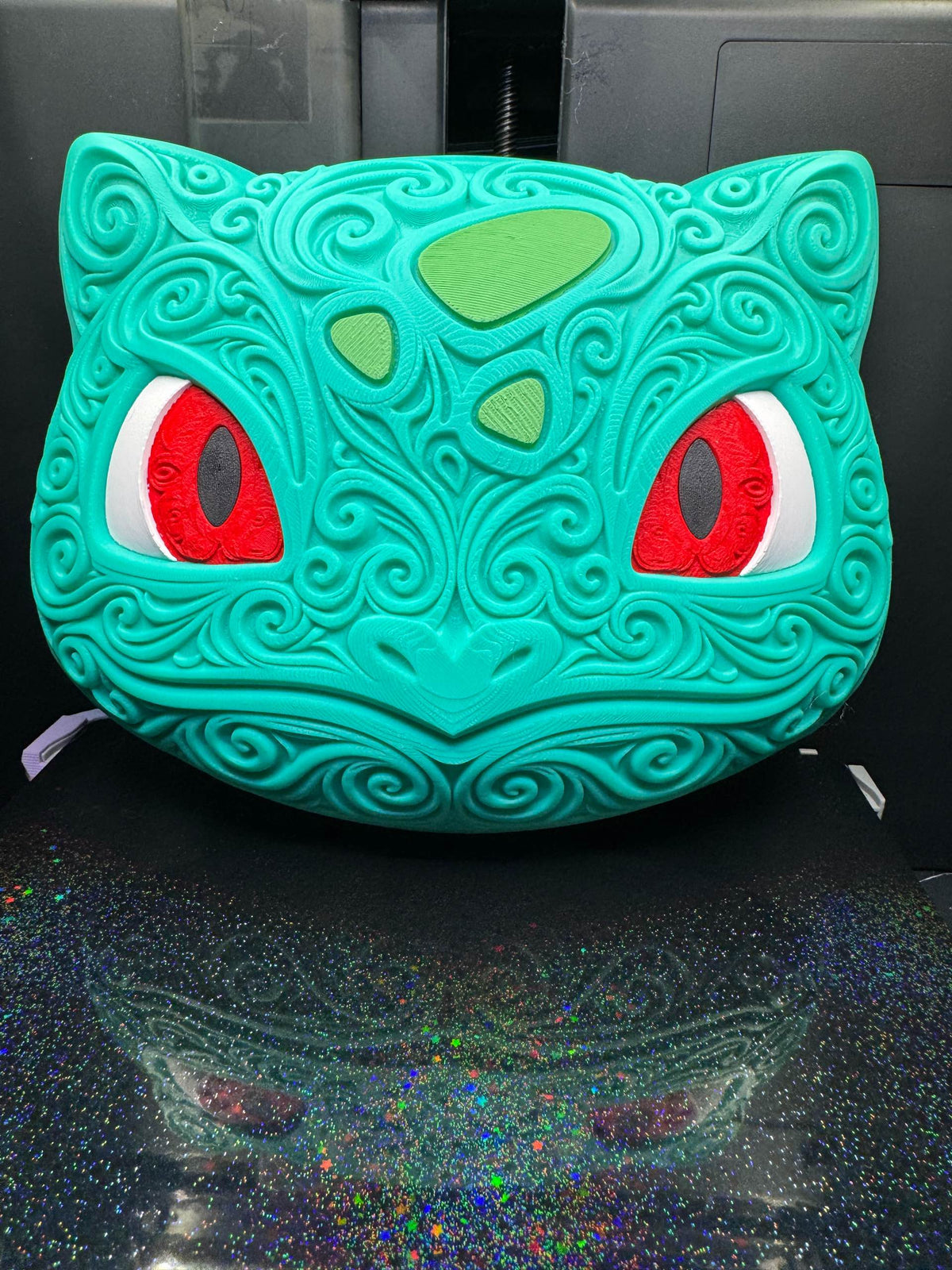 Upright front view of turquoise Bulbasaur wall art with raised scrollwork, green forehead markings, and red eyes.