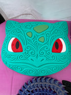 Bulbasaur wall art displayed on a pink surface, showing its ornate face and contrasting red, white, and black eyes.