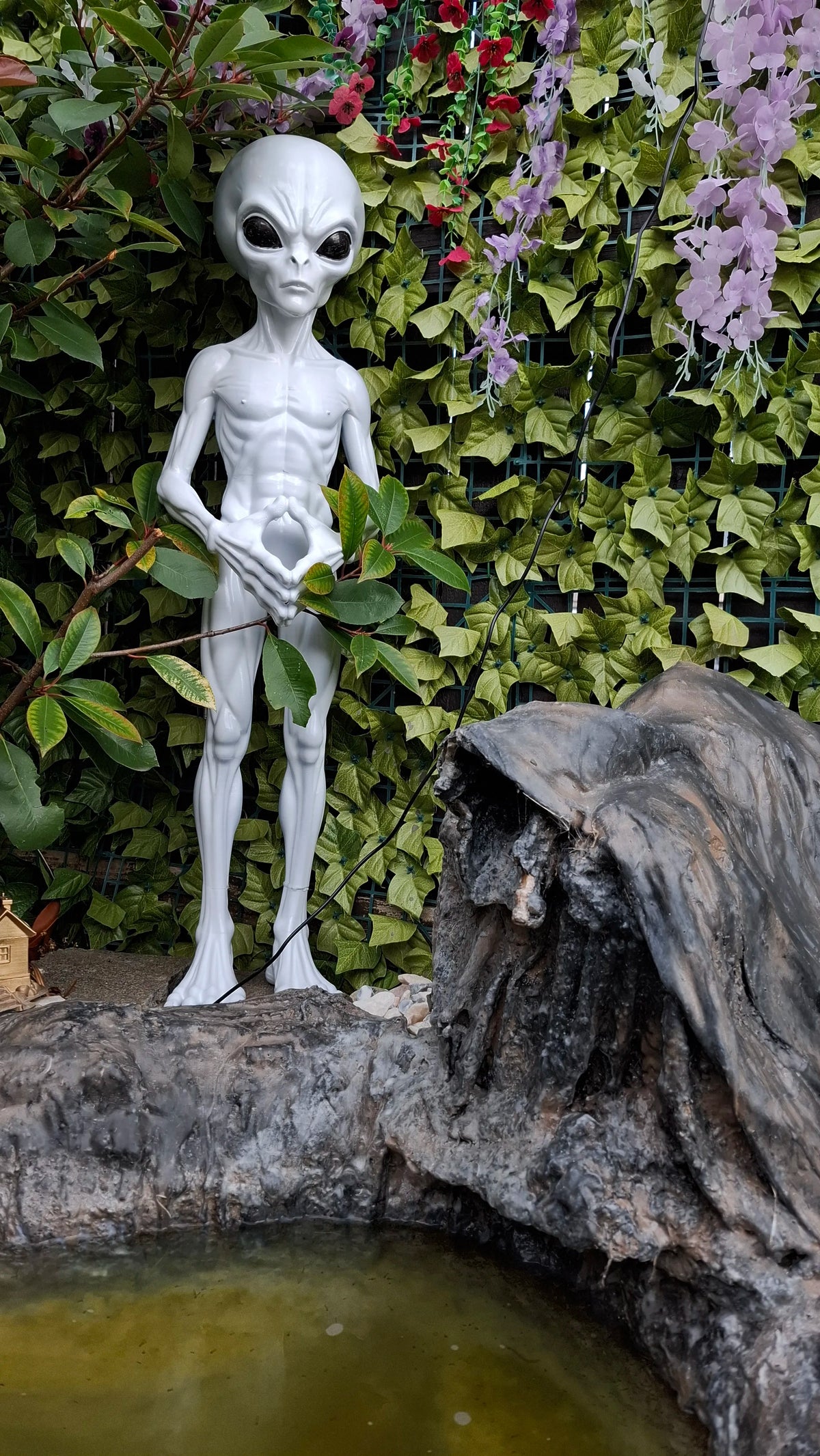Silver-gray alien statue displayed among green leaves and flowers beside a rock pond