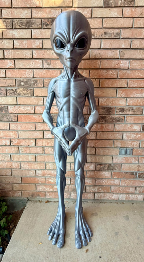 Front view of a silver-gray 3D-printed alien statue standing against a brick wall