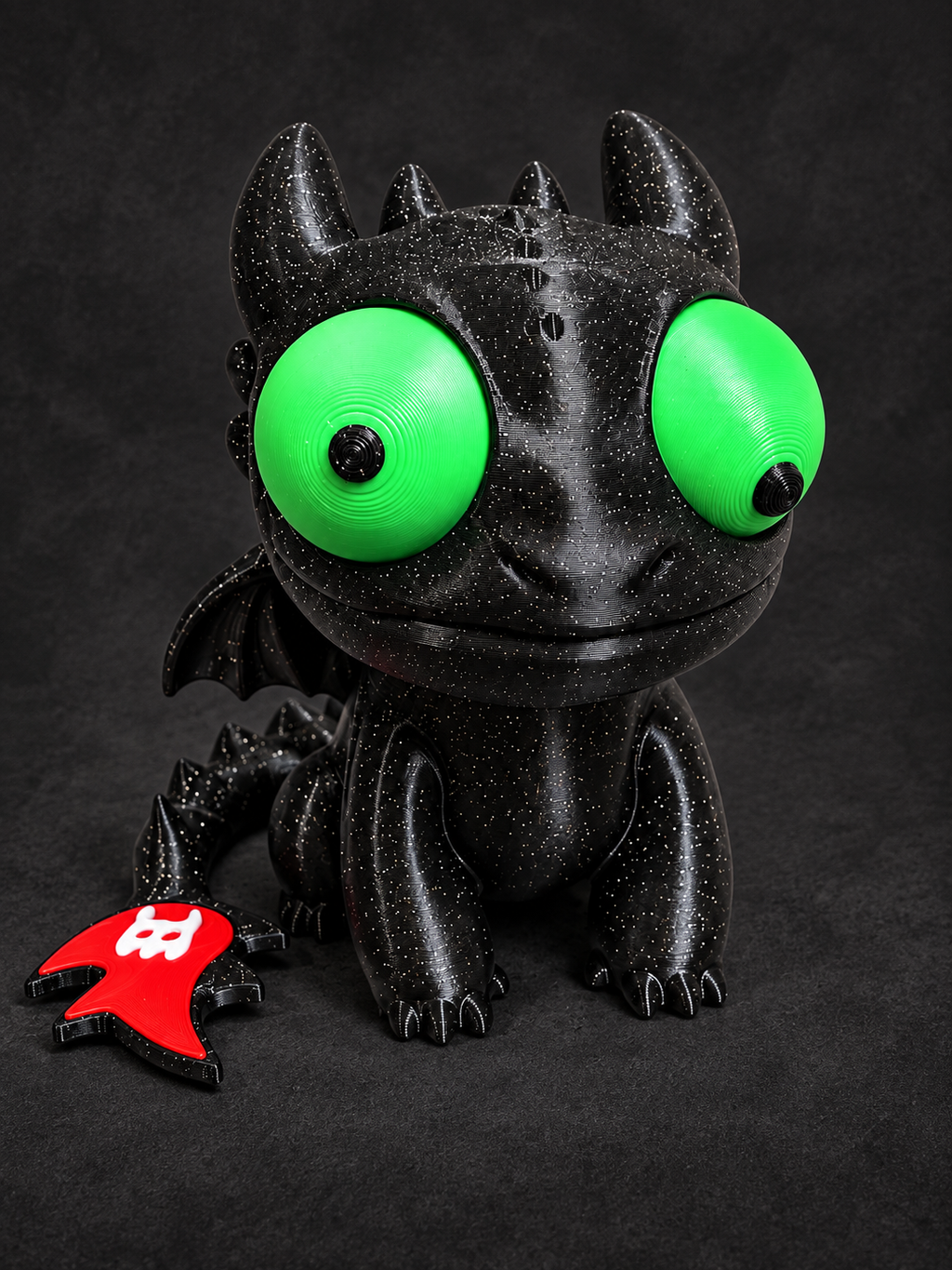 Front view of black speckled Goofy-Eyed Toothless with large movable green eyes, small wings, and a red and white tail fin on a charcoal background.