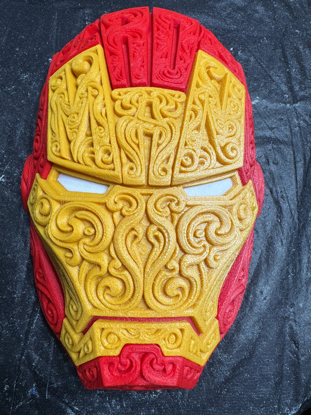 Close front view of the red and gold 3D-printed Iron Man wall piece showing raised scrollwork and pale eyes.