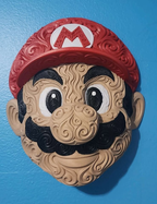 Front view of Mario wall art mounted on a blue wall, showing a red M cap, white eyes, and ornate tan and black details.