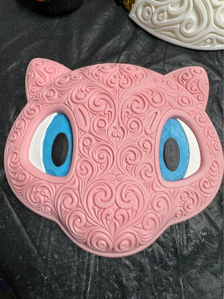 Pink Mew wall art with ornate raised filigree, blue and white eyes, and black pupils on a dark work surface.