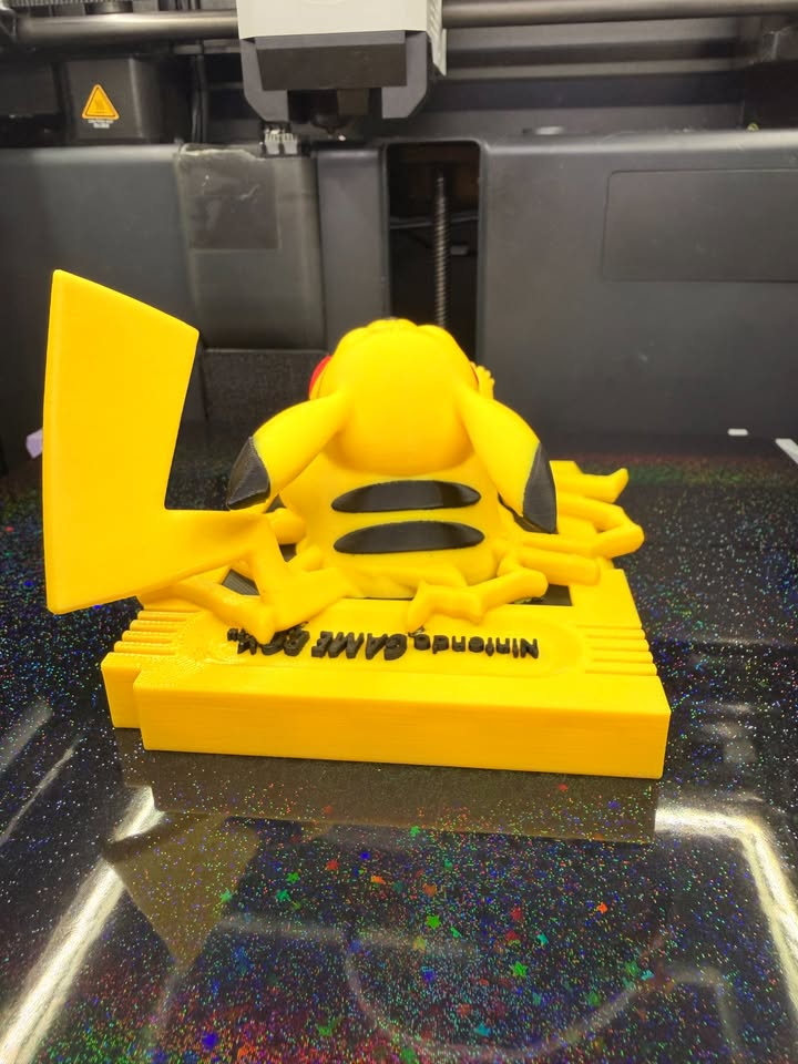 View over Pikachu's back showing black stripes, black ear tips, a yellow tail, and lettering on the cartridge-style wall plaque.