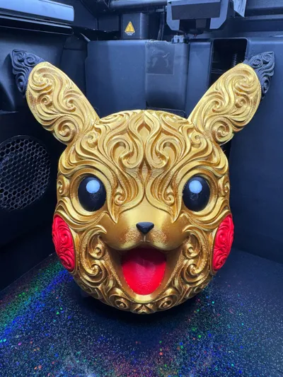 Front of gold Pikachu head wall art with ornate scrollwork, black eyes, red cheeks, and a red tongue on a 3D printer bed.