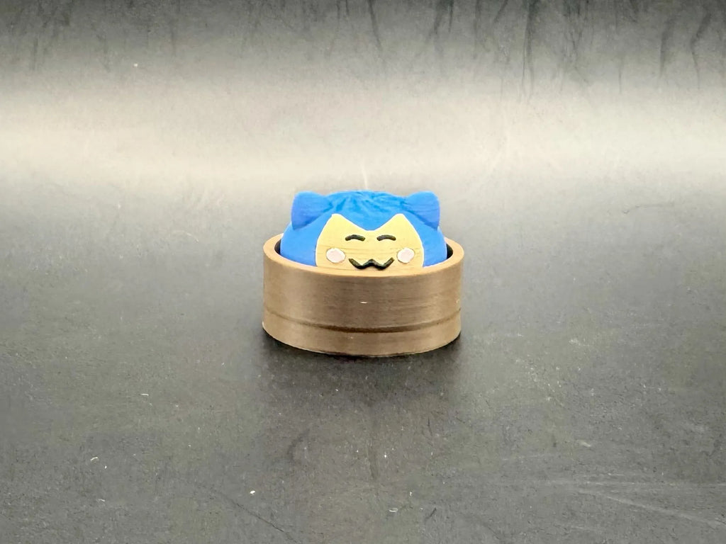 Blue Snorlax dumpling clicker with a cream face and closed eyes in a brown steamer-style base.