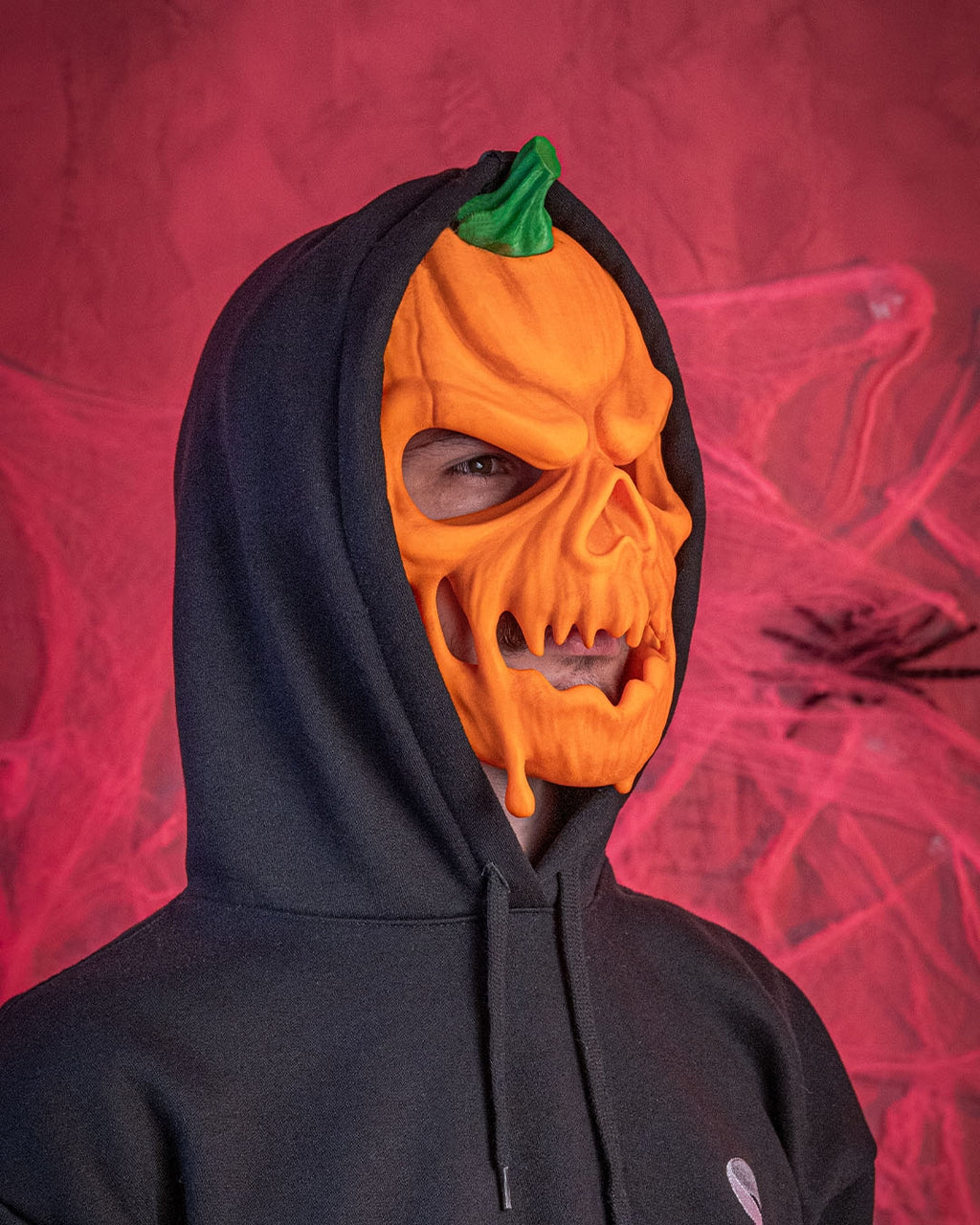 Angled fit example of the pumpkin skull Halloween mask worn with a black hood against a red background.