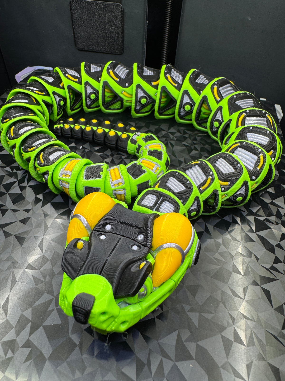 Overhead angled view of the coiled green, black, yellow, and silver robotic rattlesnake showing flexible body joints.
