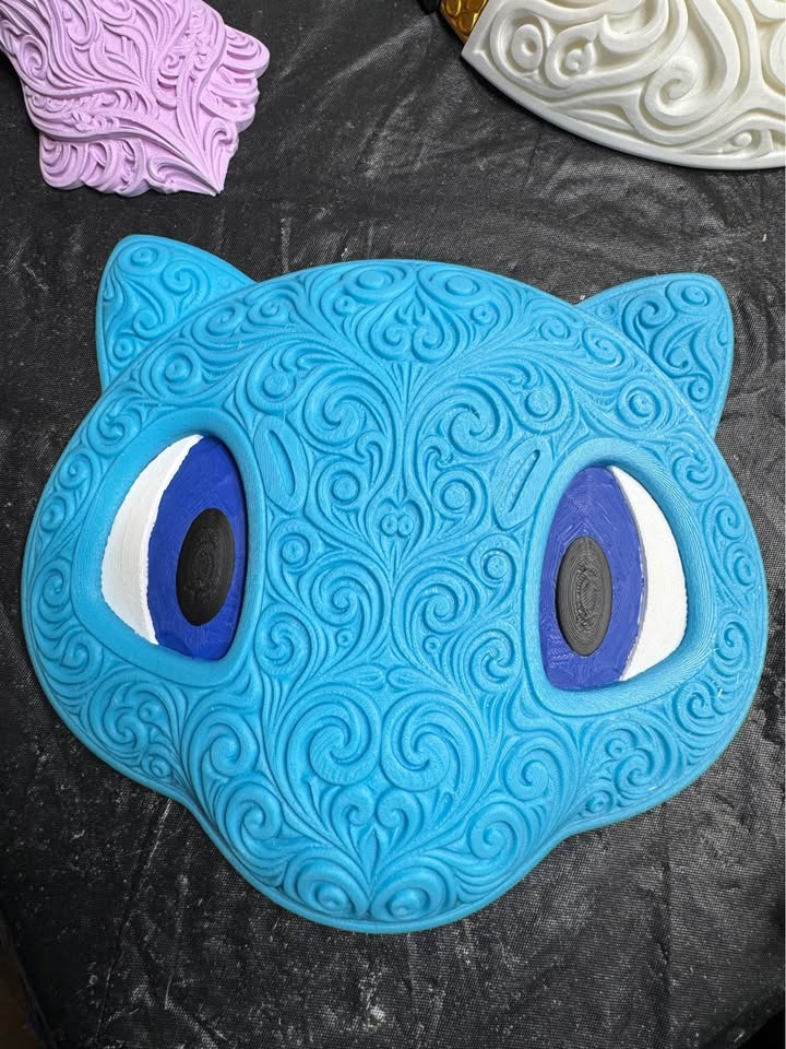 Blue Shiny Mew wall art with raised ornate filigree, purple and white eyes, and black pupils on a dark work surface.