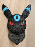 Black Shiny Umbreon wall art with raised filigree, blue forehead and ear rings, and red eyes against a wood background.