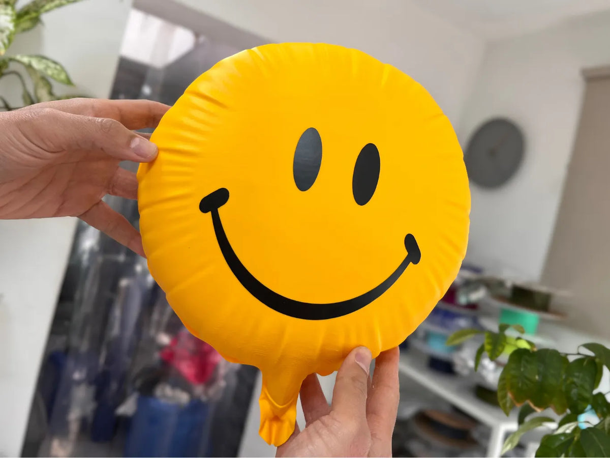 Yellow 3D printed smiley balloon with black eyes and smile, held upright to show its sculpted balloon edge.