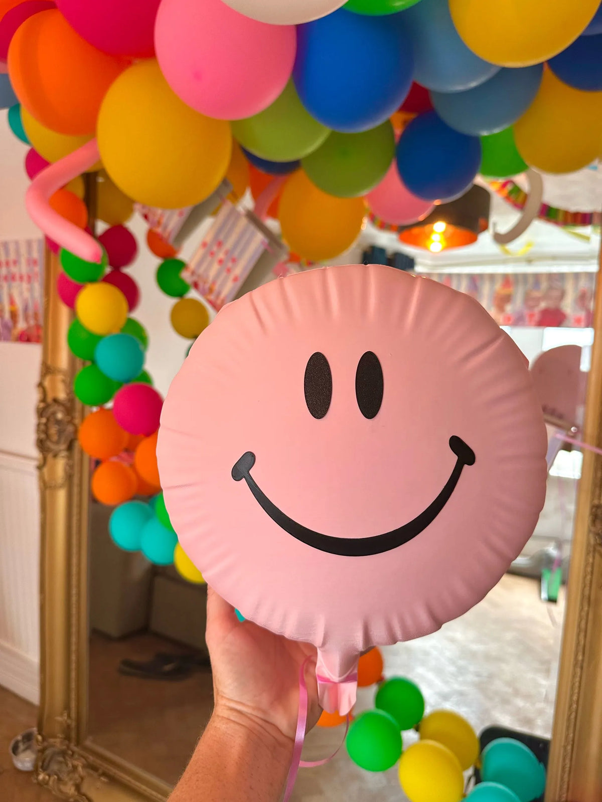 Pink smiley balloon with black eyes and smile and pink ribbon, shown among party balloons.