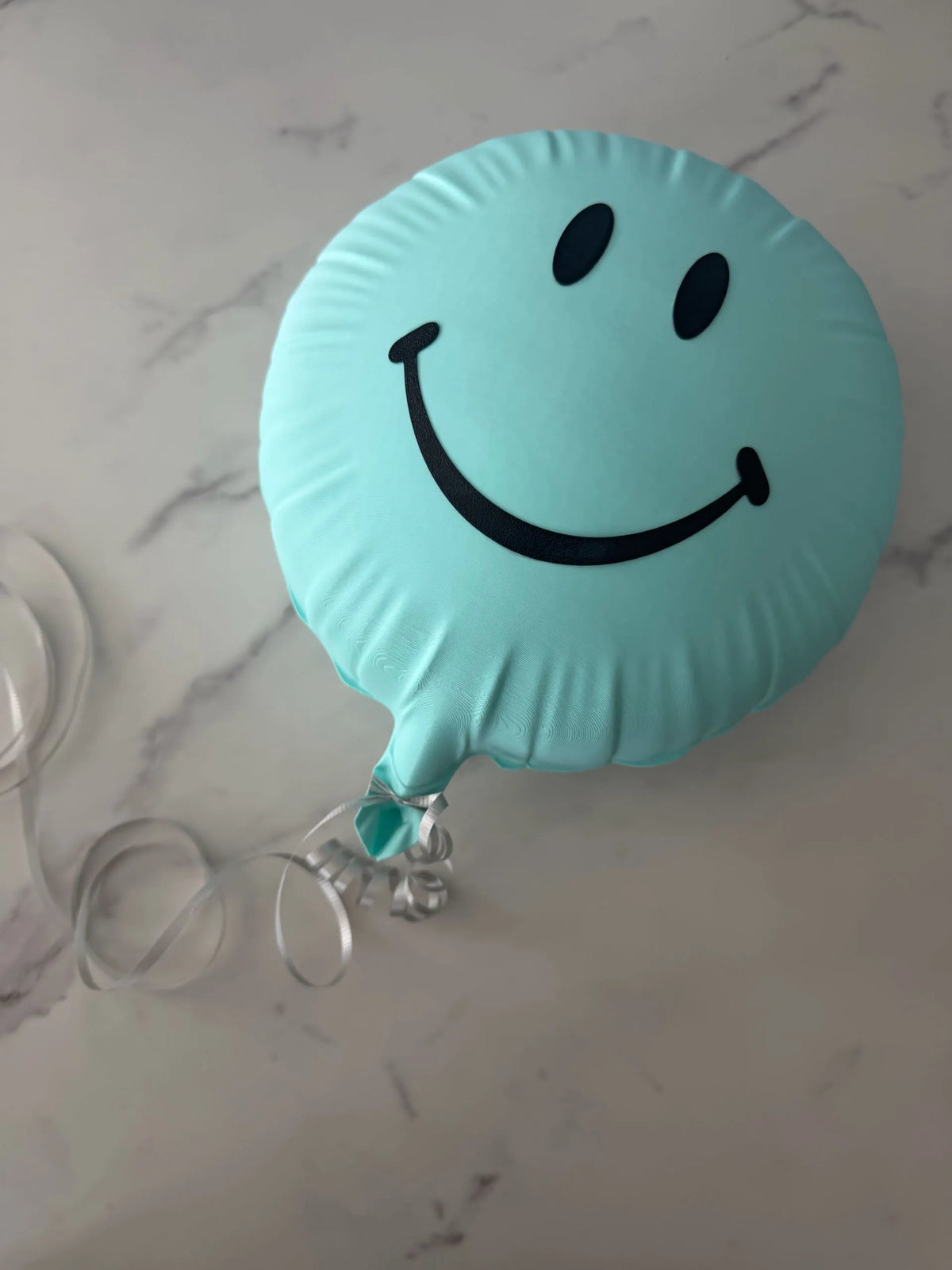 Mint-colored smiley balloon with black face and silver curling ribbon on a pale patterned surface.