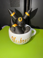Sleeping black Umbreon with gold rings inside a white teacup with gold Umbreon lettering, shown from the front detail view.