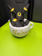 Sleeping black Umbreon with gold rings inside a white teacup with gold Umbreon lettering, shown from the front view.