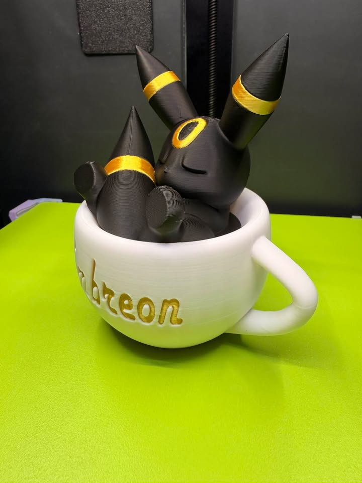 Sleeping black Umbreon with gold rings inside a white teacup with gold Umbreon lettering, shown from the side view.